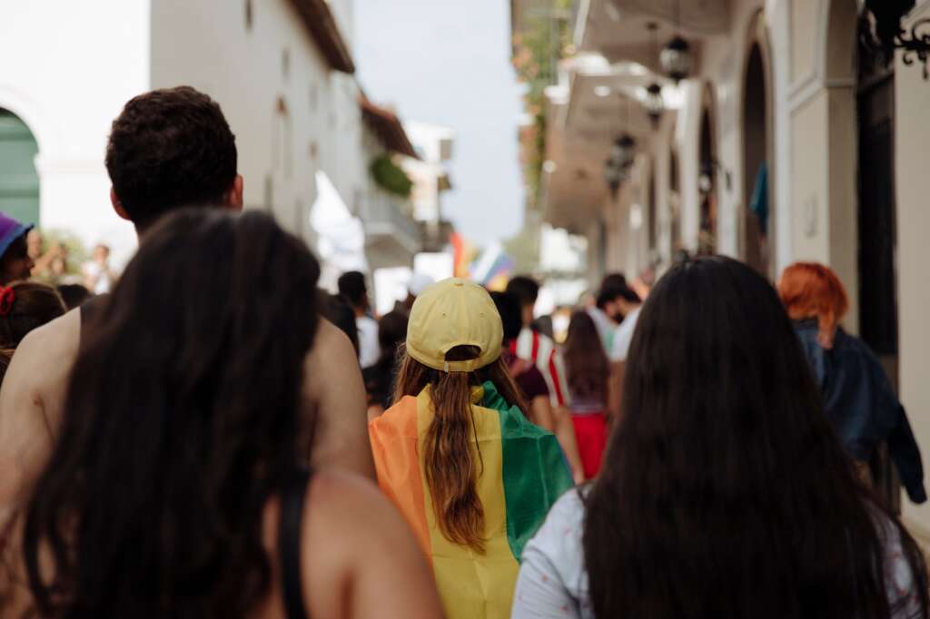 A woman wearing a yellow hat wearing a rainbow pride flag in the middle of a crowd during the lgbtq+